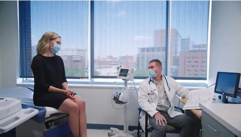 A healthcare professional in a white coat and mask discusses clinical services with a patient in a medical examination room, with diagnostic equipment nearby.