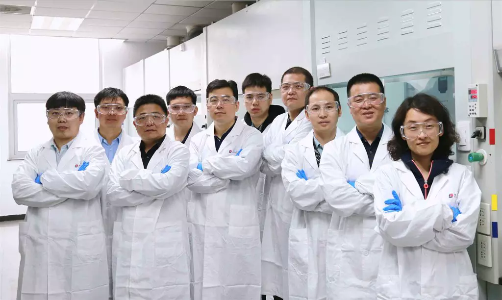 A group of 10 scientists in white lab coats and safety goggles stand with their arms crossed in a laboratory, smiling confidently.
