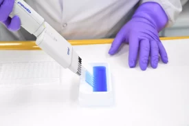 A scientist in purple gloves uses a multi-channel pipette to transfer a blue liquid into a well plate, possibly for integrated radiolabelled sciences research. The setup suggests precision handling of radioactive compounds for advanced assays.