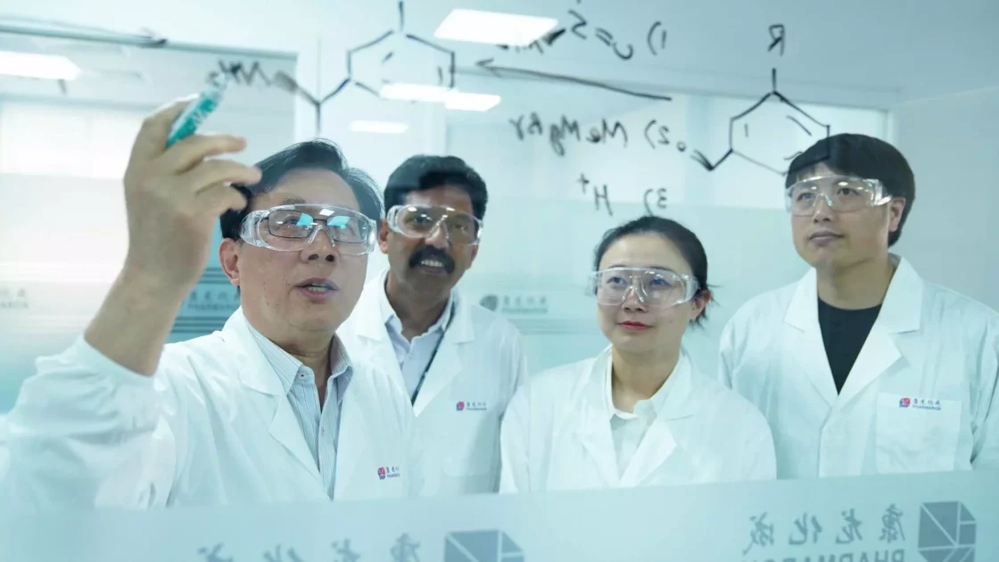 A group of four scientists in lab coats and safety glasses stand behind a glass wall with chemical formulas written on it. One scientist is writing on the glass with a marker while the others observe, focusing on bioanalytical development as they work collaboratively in the laboratory.