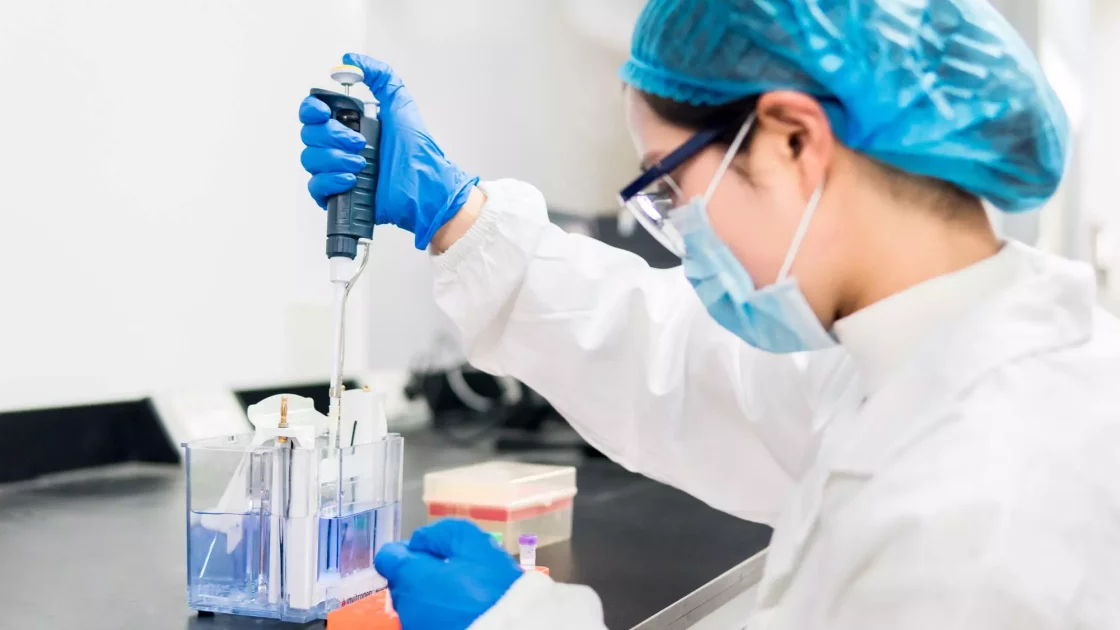 A scientific expert in protective gear uses a pipette in a laboratory to conduct an in vitro disease biology experiment involving liquid samples.