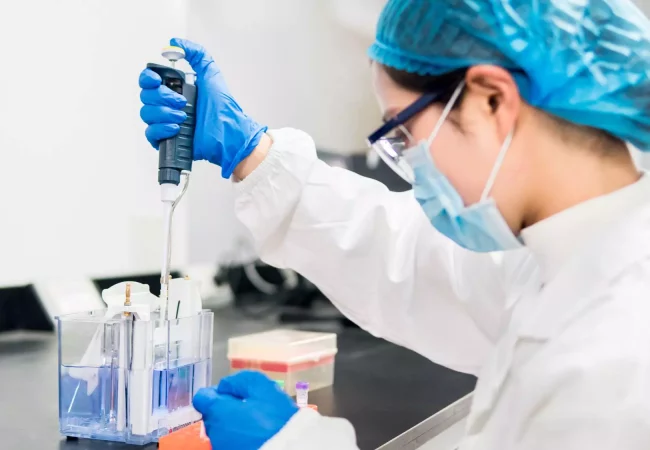 A scientific expert in protective gear uses a pipette in a laboratory to conduct an in vitro disease biology experiment involving liquid samples.
