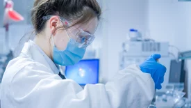 Scientist in lab coat, gloves, and safety glasses handling a sample vial during CGT in vivo pharmacology preclinical studies in a bright laboratory setting