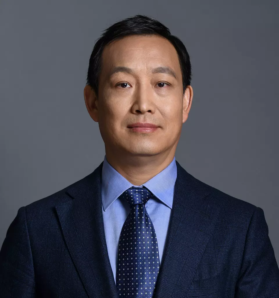 Larry Lou, EMBA, M.Eng - A man with short dark hair is seen in a professional headshot. He is wearing a dark blue suit jacket, a light blue dress shirt, and a navy blue tie with white polka dots. His arms are crossed and he is looking confidently at the camera against a plain gray background, exemplifying absolute professionalism.
