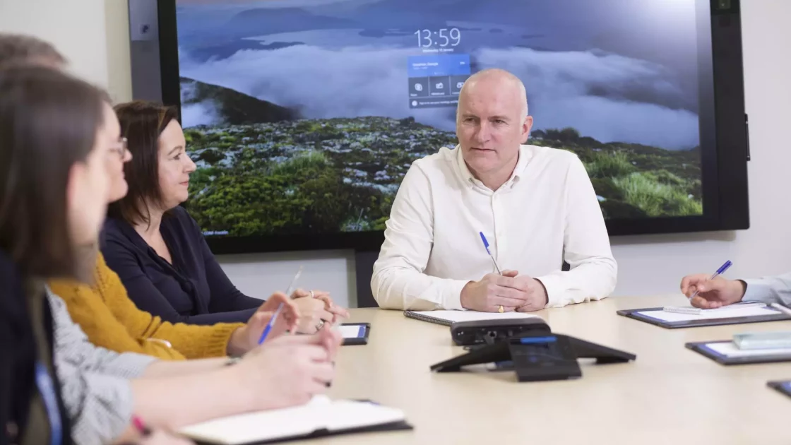 A team discusses regulatory dossier authoring and specification justification around a conference table, with notes and devices in hand.