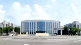 A modern, expansive office building with a sleek glass facade stands under a blue sky with scattered clouds. The building is surrounded by green trees, and a well-maintained garden area is in front. Several cars are parked on the street in front of the building that houses cutting-edge preclinical ADME studies labs.
