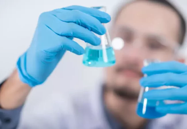 A scientist in blue gloves carefully examines two beakers containing blue liquid, focusing on protein binding analysis.