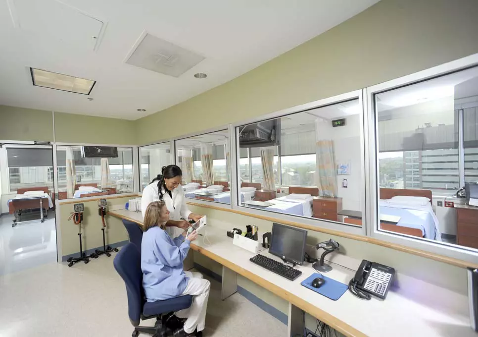 Two healthcare professionals discuss patient data in a control room overlooking multiple hospital beds. The setting indicates integrated clinical development services, with a focus on monitoring and coordinating clinical trials or patient care in a controlled environment.