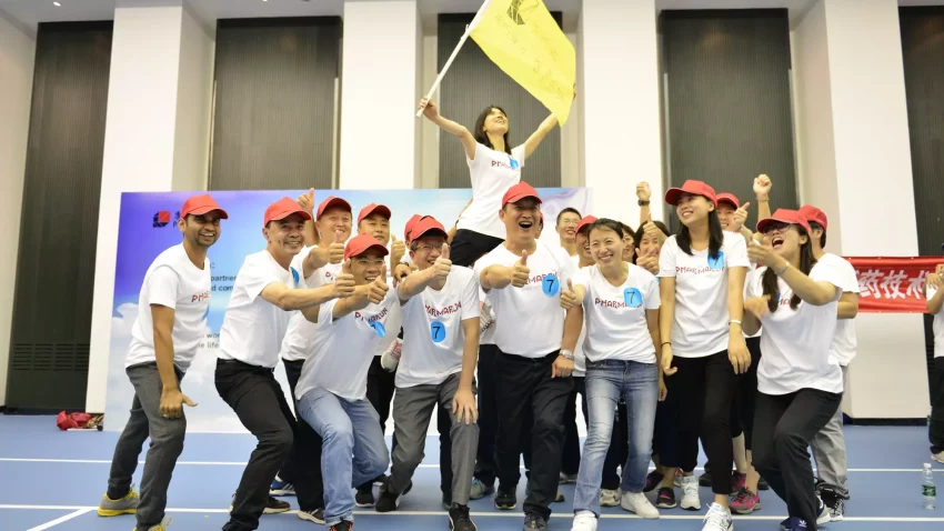 A jubilant Pharmaron team poses together, wearing matching shirts and red caps, celebrating with thumbs-up gestures, one member holding a yellow flag.