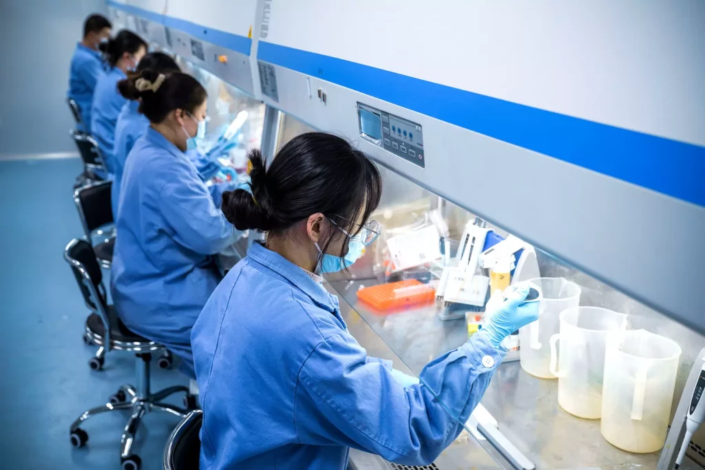 Scientists in blue lab coats and face masks are working in a row at laboratory benches, focusing on genetic toxicology. They use pipettes, containers, and other lab equipment under a sterile hood. The lab environment appears clean and organized.