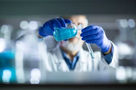 A scientist wearing blue gloves carefully pours blue liquid from a flask into a test tube in a laboratory setting. The background is blurred, emphasizing the precise handling of chemicals. in radiosynthesis and radiolabelling services for ADME