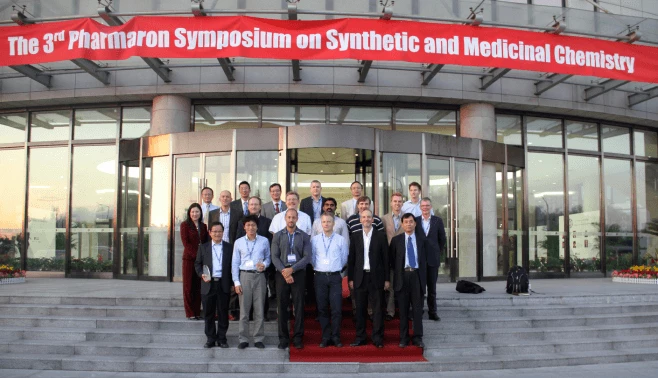 A group of people, dressed in business attire, stands on the steps outside a building with a glass facade. Above them, a red banner reads "The 3rd Pharmaron Symposium on Synthetic and Medicinal Chemistry." The event appears well-organized and professional, highlighting topics like absolute bioavailability and preclinical ADME studies.