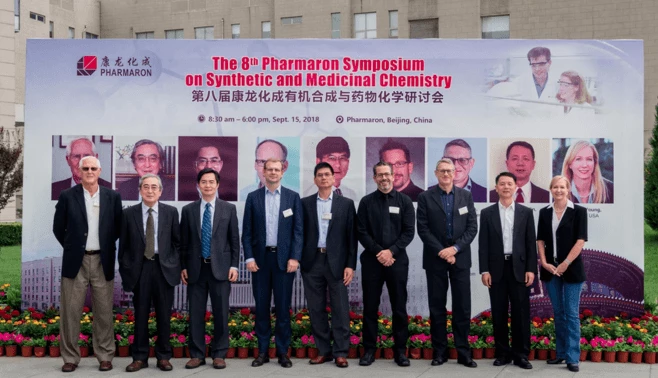 A group of ten people standing in front of a banner at the 8th Pharmaron Symposium on Synthetic and Medicinal Chemistry in Beijing, China. The banner, showcasing images of various scientists involved in Radiolabelled ADME studies, includes text in both English and Chinese. The people are smiling at the camera.