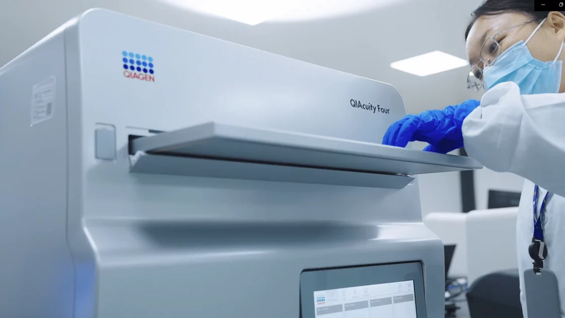 A scientist wearing a mask and gloves operates a QIAcuity Four digital PCR instrument, carefully placing a sample for cell line engineering analysis.