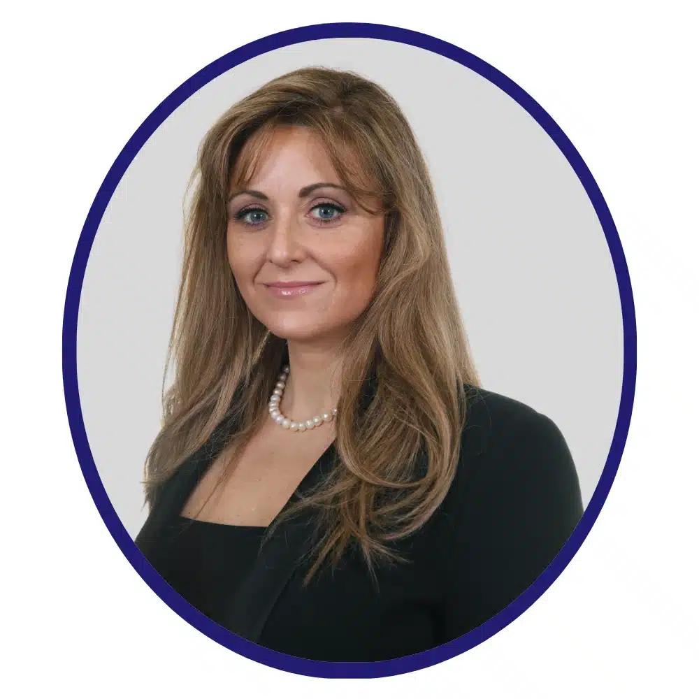 Professional headshot of a smiling woman with long, wavy brown hair, wearing a black blazer, pearl necklace, and a black top, framed within a circular border.