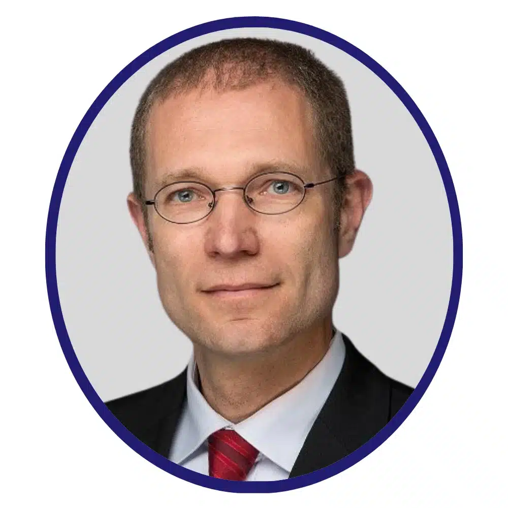 Professional headshot of a man with glasses, wearing a black suit, white shirt, and red tie, framed in a circular border on a neutral background.
