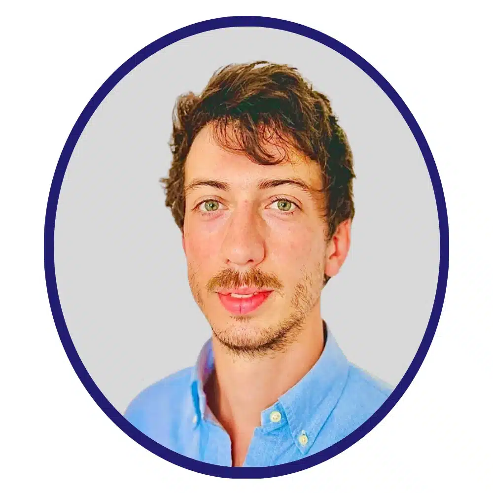 Young man with wavy brown hair and a mustache, wearing a light blue collared shirt, framed in a blue-bordered oval.