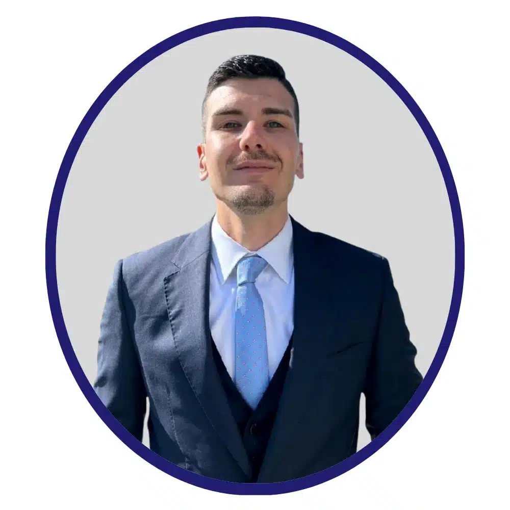 Man in a navy blue suit with a light blue tie, standing outdoors, framed in a blue-bordered oval.