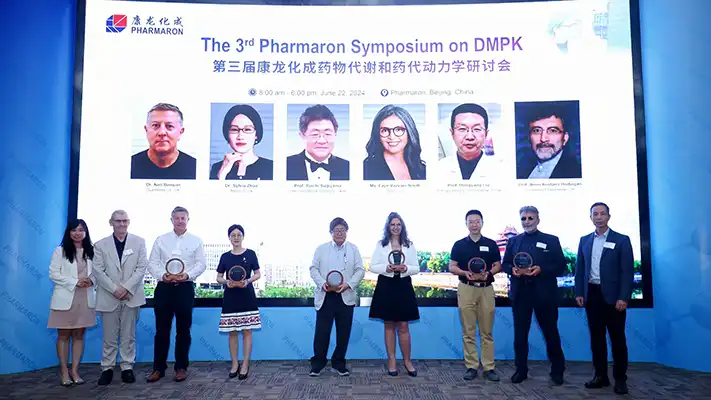 Speakers at the 3rd Pharmaron Symposium on DMPK stand on stage holding plaques, with a backdrop displaying their photos and names.