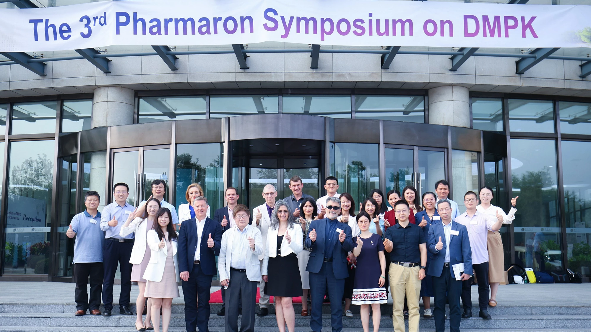 The management team at the 3rd Pharmaron Symposium on DMPK poses in front of the venue, smiling and giving thumbs up beneath the event banner.