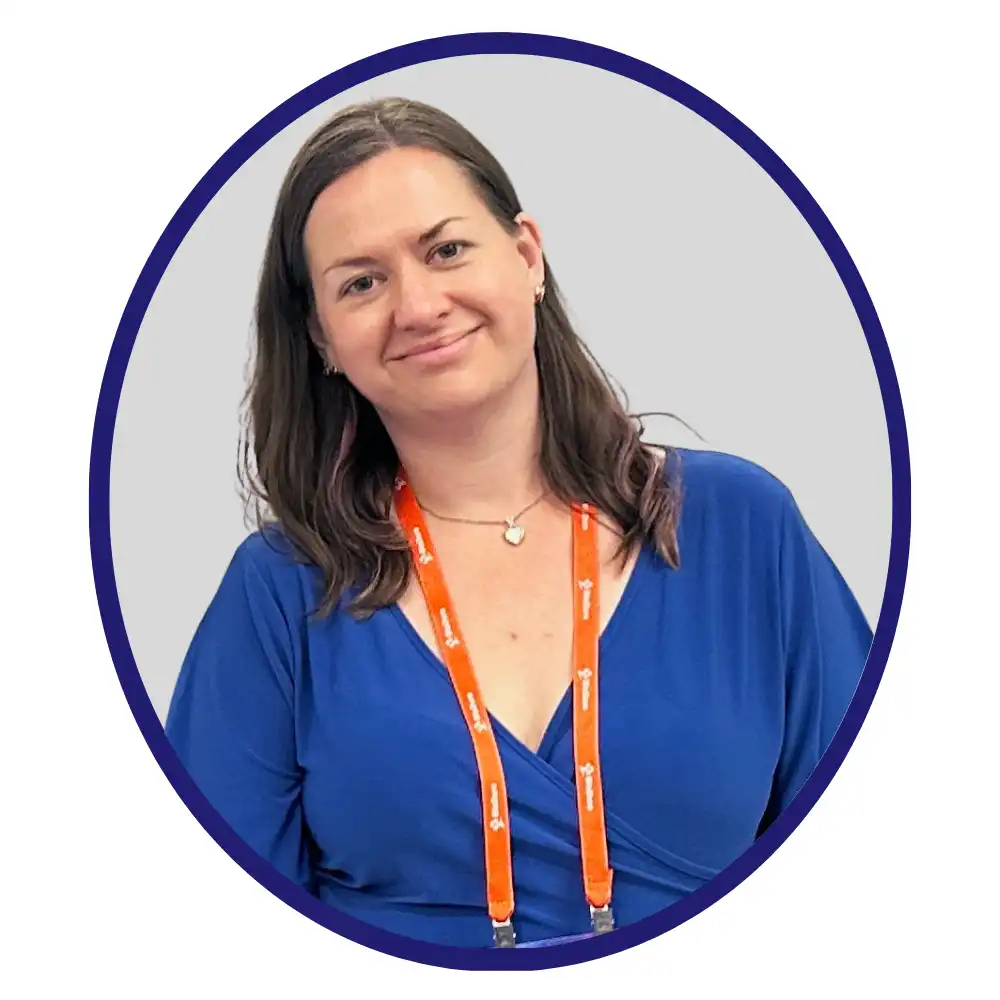 Smiling scientist in a blue blouse wearing an orange conference lanyard, framed in a dark blue oval on a light gray background.