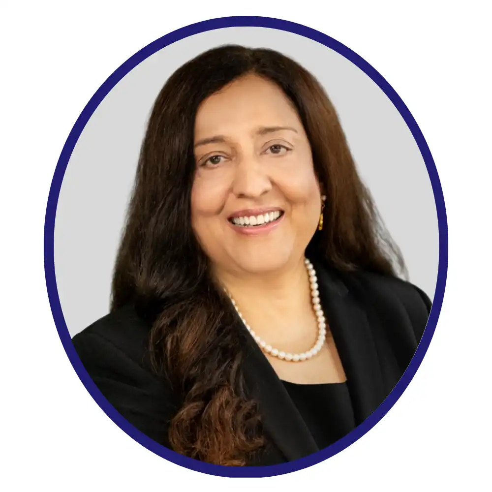 Smiling woman with long dark hair wearing a black blazer, pearl necklace, and gold earrings, framed in a blue oval on a gray background.