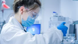 Scientist in safety goggles and mask inspecting a sample vial in a modern lab for CGT bioanalysis services