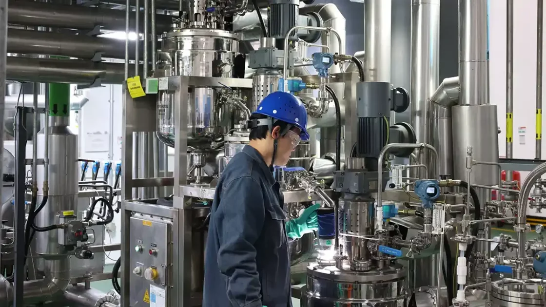 Scientist in hard hat and gloves operating large-scale stainless steel reactor vessels in a continuous flow chemistry manufacturing facility.