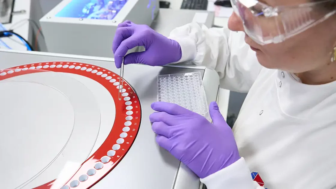 Scientist in PPE using tweezers to load a sample into a numbered carousel while holding a 96-well plate